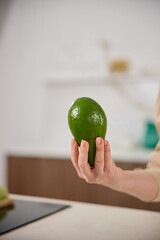 A large avocado in a woman's hand in the interiors of a bright and spacious kitchen