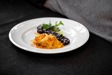 A close-up, elevated-angle shot of a gourmet seafood dish featuring tender squid rings, a vibrant orange sauce, and a side of dark black rice on a white plate in a restaurant setting.