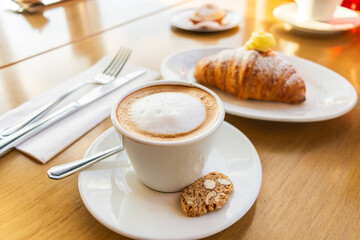 Primer plano de taza de capuchino espumoso y galleta de almendras en mesa de madera, con croissant relleno de crema desenfocado al fondo. Desayuno en cafetería soleada.
