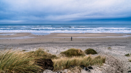 Solitary Figure Walks Along Quiet Beach Under Cloudy Sky With Dunes and Grass, Kandestederne, Denmark