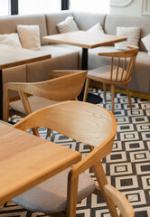 Cozy modern cafe interior with light wood chairs and tables, featuring a built-in beige banquette sofa with pillows. The floor has a black and white geometric tile pattern. Focus is on the foreground.