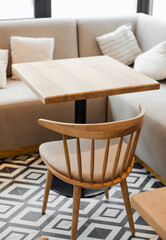 Cozy modern cafe interior. Light wood table and a matching chair against a beige banquette sofa with pillows. The floor has a black and white geometric tile pattern. 