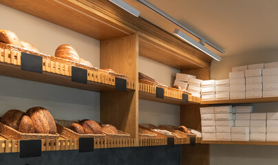 Modern bakery display with artisanal bread loaves sit in wicker baskets on wooden shelves to the left. To the right, tall stacks of plain white takeout boxes are stored on shelves.