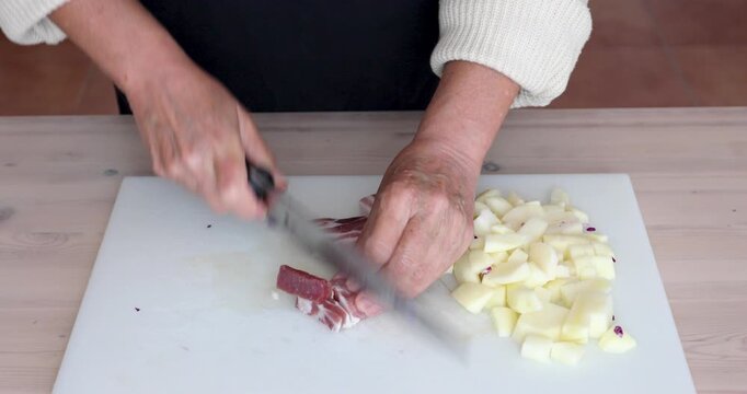 Close-up of hands cutting pancetta into small cubes on a white plastic cutting board. On the side of the board, a small pile of diced potatoes is already prepared. No audio. Food preparation concept.