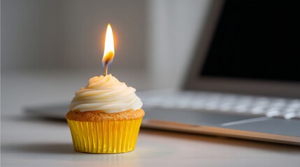 Cupcake with lit candle on a desk near a laptop, symbolizing a remote work birthday celebration, anniversary, or special event