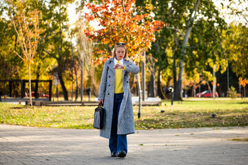 Urban businesswoman with sunglasses in stylish autumn coat checking the time on her wristwatch while walking with a laptop bag through the city park during sunny day.