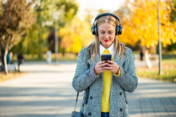 Stylish businesswoman in warm coat using wireless headphones and smartphone while standing in the city park during sunny autumn day.
