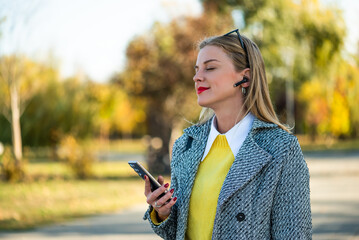 Close up portrait of beautiful urban businesswoman in autumn coat listening to music through earphones and using smartphone while standing in the city park on a sunny day.	