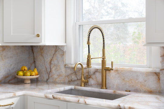 A gold kitchen faucet detail with white cabinets, stainless steel sink, and granite countertops.