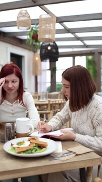 Vertical screen nutritionist reviews the clients food diary in a cafe while the client reacts with discomfort creating an awkward moment as the expert tries to explain her habits and next steps