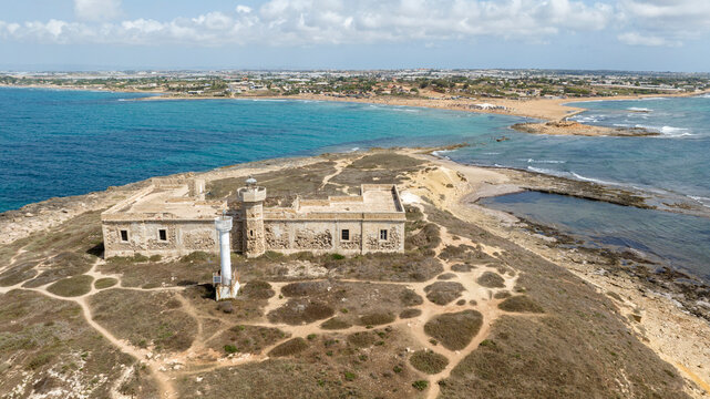 Aerial close-up of Isola delle Correnti, near Portopalo in Sicily, Italy. On the rocky island are the remains and ruins of an old building, once used as a barracks, and a lighthouse. Sunny summer day.