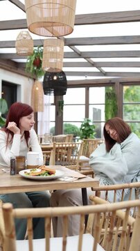 Vertical screen two friends sit in a cafe as one talks with lively energy unaware that the other has quietly fallen asleep wrapped in a blanket turning their chat into a funny cozy moment