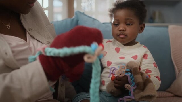 Young African American woman wearing glove puppet playing with her toddler daughter while sitting together on sofa