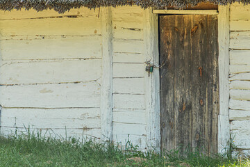 Old wooden building with thatched roof, located in an ancient Central European village Natural setting with vegetation and uneven ground Large central door with padlock, dark in color No people pr
