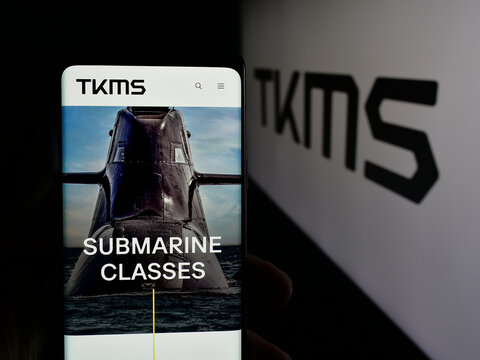 Stuttgart, Germany - 11-20-2025: Person holding cellphone with webpage of company ThyssenKrupp Marine Systems (TKMS) on screen in front of logo. Focus on center of phone display.