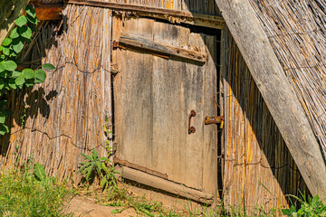 Rustic, thatched-roof wooden hut in a rural Central European setting, open door revealing dark interior, patches of greenery nearby, naturalistic perspective, daytime with clear sky, high contrast st