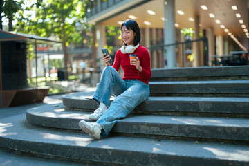Asian woman smiling using phone on city stairs enjoying coffee