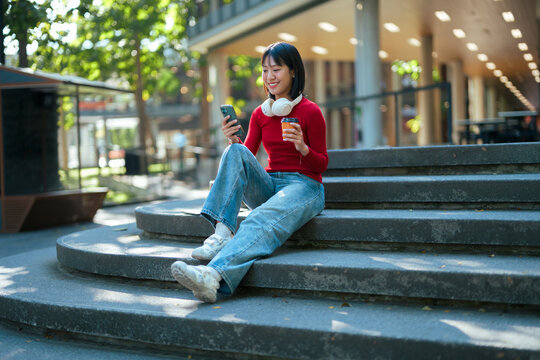 Asian woman smiling using phone on city stairs enjoying coffee