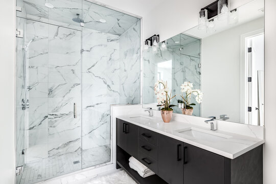 A beautiful bathroom with a dark vanity and white granite counter top. A plant and towels sit on the vanity. The shower is lined with marble tiles. Lights off.