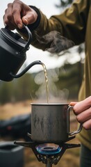 Person pouring steaming hot coffee from kettle into metal mug on portable gas stove outdoors