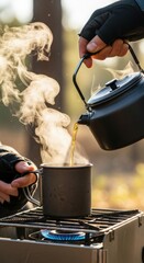 Gloved hands pouring steaming hot drink into metal mug on portable camping stove outdoor adventure