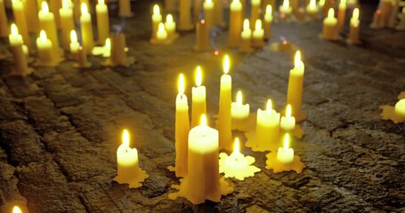 Flickering candlelight creates a warm ambiance in a dimly lit area. The candles of various heights are scattered on a textured surface, casting soft shadows. - Powered by Adobe