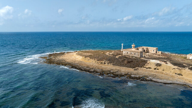 Aerial close-up of Isola delle Correnti on Mediterranean Sea, near Portopalo in Sicily, Italy. On the island are the remains and ruins of an old building, once used as a barracks, and a lighthouse.
