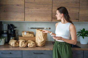 Young woman checks grocery delivery using a mobile app while standing in the kitchen. Concept of app-based meal kit subscription and digital grocery planning for home cooking.