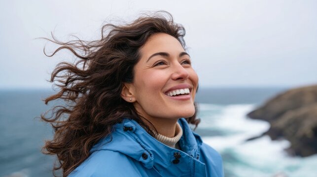 Woman smiling at camera, standing outdoors near ocean, wearing blue jacket and backpack, with wind blowing hair. - Powered by Adobe