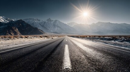 Open highway stretches toward distant, snow-capped mountain range under bright sun flare