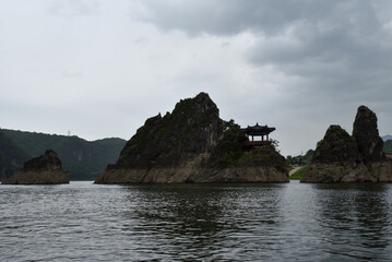 A rocky island with a small building on it. The island is surrounded by water. The water is calm and the sky is cloudy