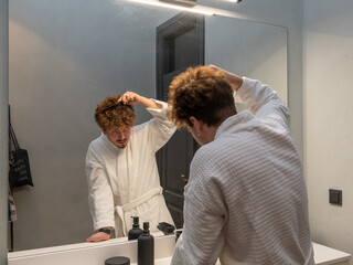Man in a white bathrobe combing his hair in the bathroom in front of the mirror