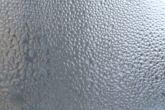 Macro close-up of condensation droplets on a smooth glass surface. A dense pattern of tiny water drops creates a textured abstract background with cool tones and soft natural light.