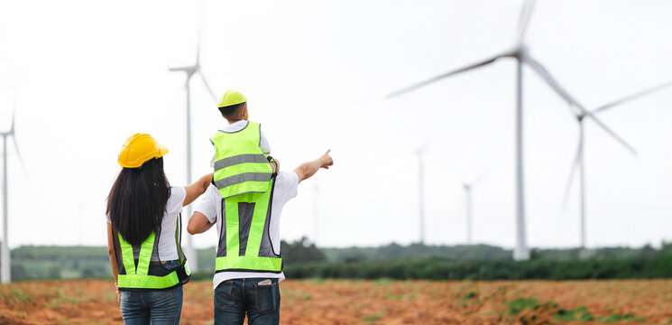 Back view Asian Engineer family in field with wind turbines in the background, Windmill. environmental renewable clean energy. Wind power generation. Solar panel. Windmill engineer inspection progress - Powered by Adobe