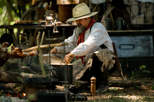 A cowboy preparing food at a rustic outdoor campfire, seasoning and stirring a steaming soup pot - Powered by Adobe