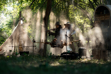 Two cowboys aiming rifles in a forest camp, with sunlight streaming through the smoke