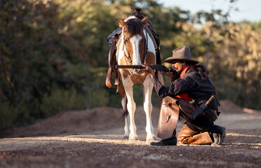 A cowboy kneeling on the ground while aiming a rifle beside his horse in a rugged outdoor setting