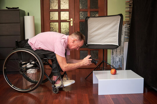 A man in a wheelchair bends over, holds a camera with both hands, and takes a picture of an apple