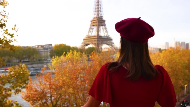 Rear view of a woman in a red dress and beret hat enjoying the view of the Eiffel Tower in Paris, France, from her balcony during a golden sunrise