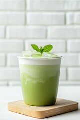 Iced matcha green tea latte with fresh mint garnish and ice cubes in a cup on a wooden coaster against a white brick wall background