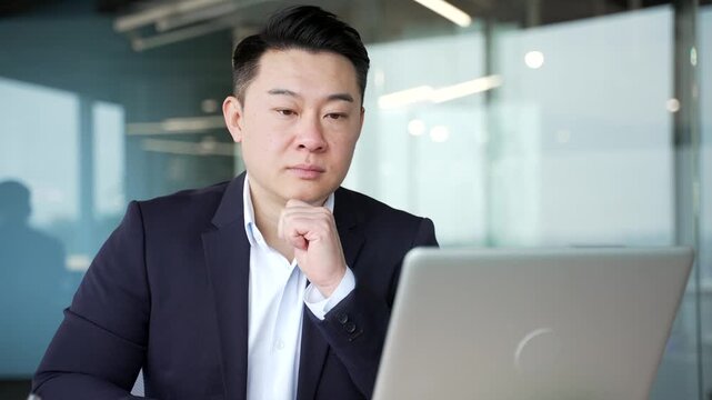 Thoughtful asian businessman working on laptop in business office. Manager thinking about problem solving at workplace. The worker is engaged in a task or is busy with a project on computer. Close up