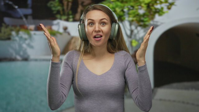Young woman wearing headphones shows surprise in an outdoor resort pool setting, capturing a fun and joyful moment.