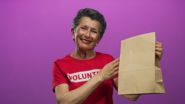 Senior woman volunteer with grey hair smiles while holding a paper bag against a vibrant pink background, symbolizing charity and food distribution.