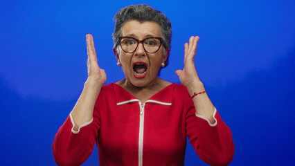 Naklejka premium Elderly woman with grey hair wearing glasses and a red top expressing surprise against a vibrant blue background.