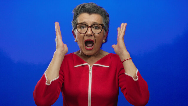 Elderly woman with grey hair wearing glasses and a red top expressing surprise against a vibrant blue background.