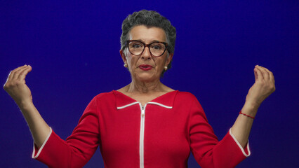 Senior woman with grey hair in red sweater meditates against a blue background with hands raised emphasizing calm and focus, illustrating mindfulness and maturity in a vibrant setting.