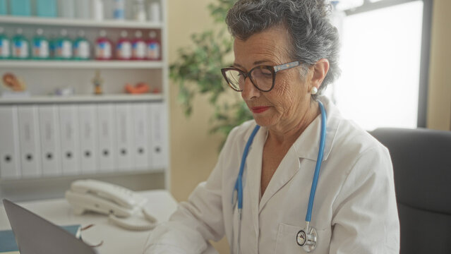 Senior female doctor with grey hair working in a modern clinic office using a laptop, surrounded by shelves of medical supplies and documents, portraying professionalism and care.
