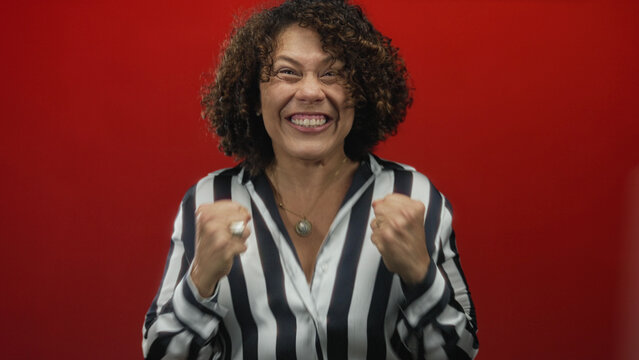 Hispanic woman wearing striped blouse clenches fists in red studio; enthusiasm empowerment triumph flourish.