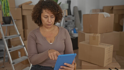 Hispanic woman tapping tablet while seated at table in building surrounded by moving boxes and packing materials; serenity.