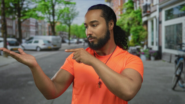Man pointing to open palm on street wearing bright orange shirt and whistle necklace near parked cars and bicycle; thoughtful reflection.